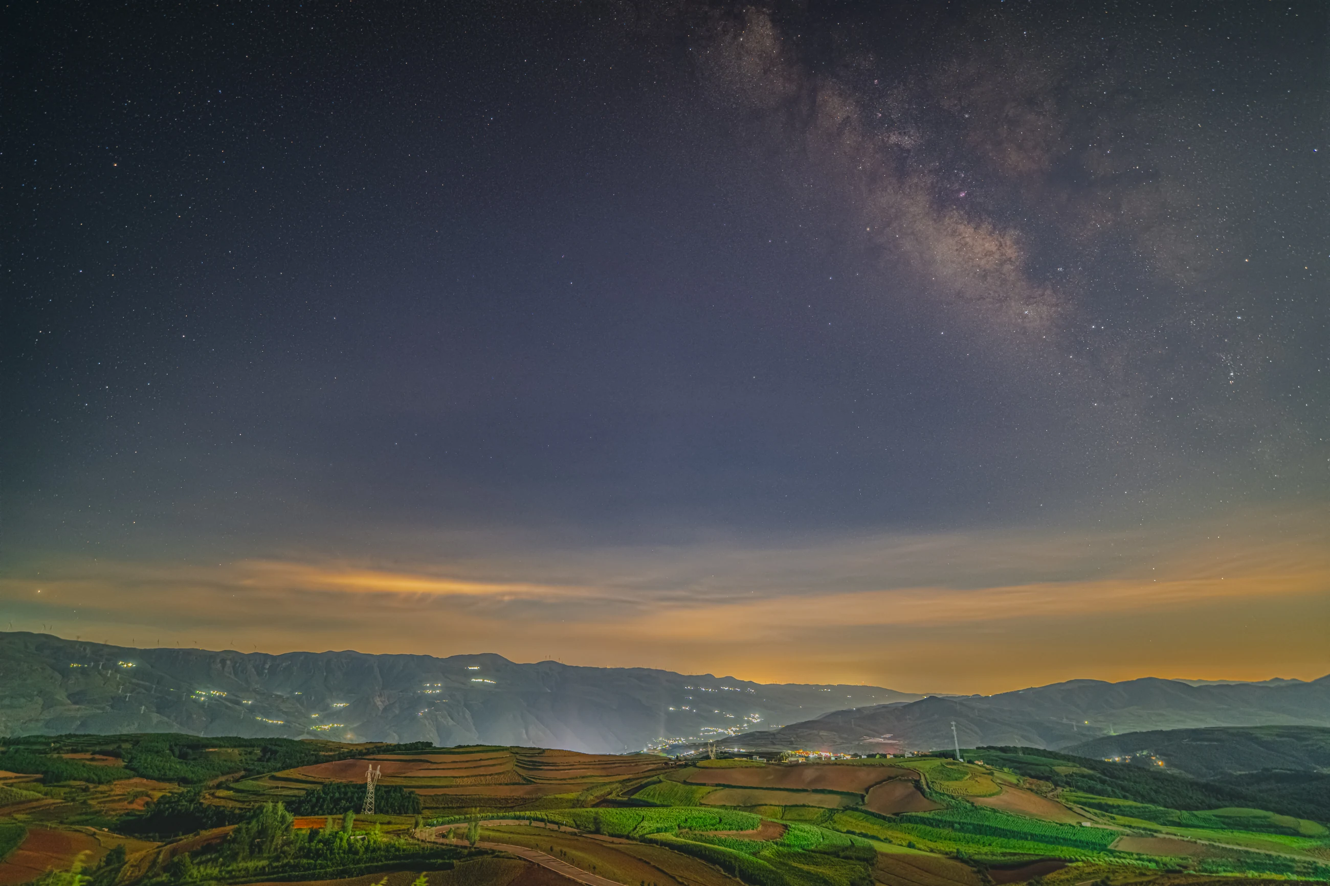 The Milky Way above layered agricultural hills and distant lights at Dongchuan Red Land.